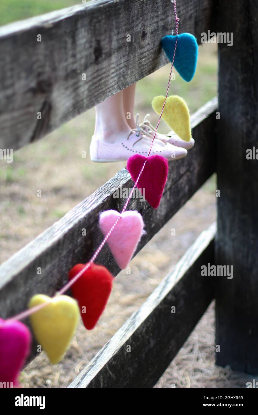 Eine regenbogenfarbene Girlande aus Herzen, die an einem Holzzaun hängen. Eine Frau mit rosa Schuhen steht auf dem Zaun Stockfoto