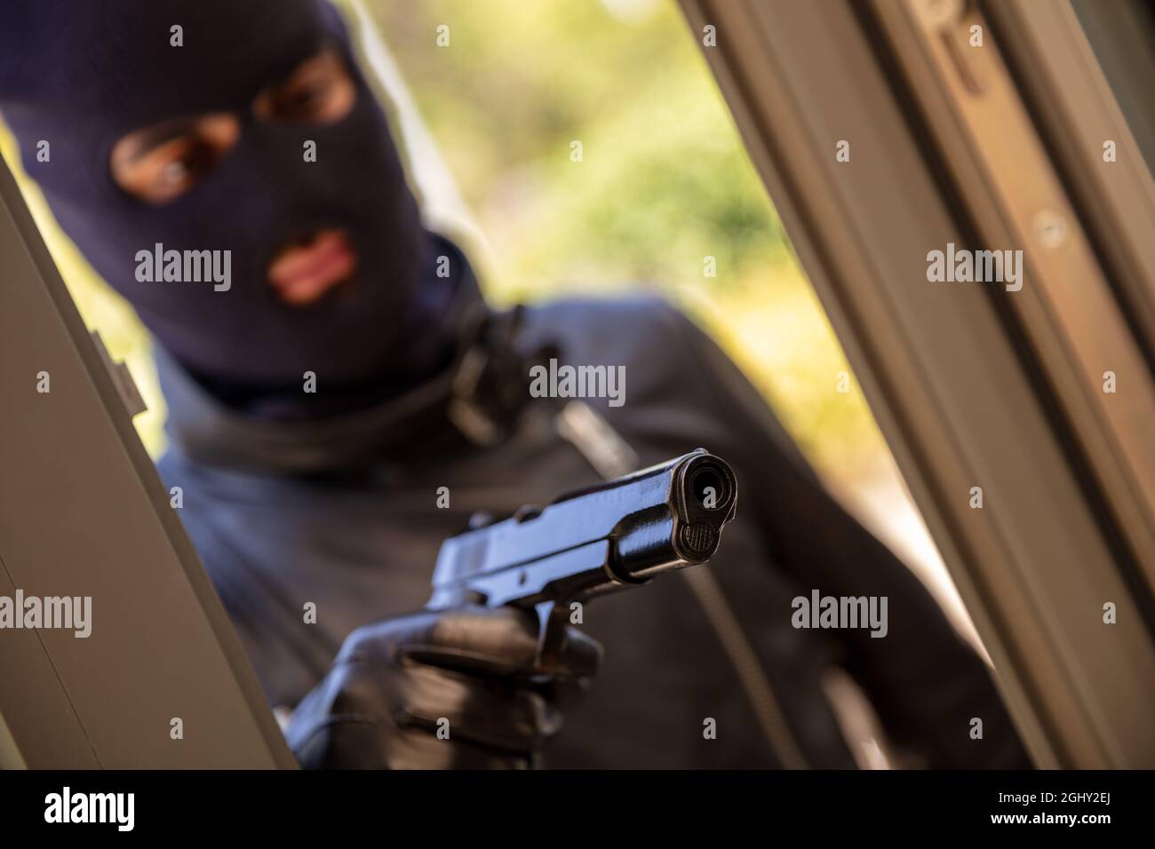 Mann in schwarzer Balaclava, der Pistole mit Lederhandschuh im Handhintergrund hält. Blur mit Kapuze bewaffneter Räuber zielt mit Waffe hinter dem Fenster. Mörder Dieb Mord Stockfoto