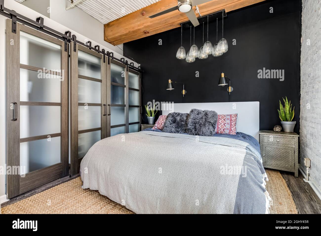 Ein Schlafzimmer in einer Loft-Wohnung mit einer freiliegenden Holzdecke, Glasschiebetüren zu einem Schrank, einer schwarzen Akzentwand und einem schicken Glaslicht, das darüber hängt. Stockfoto