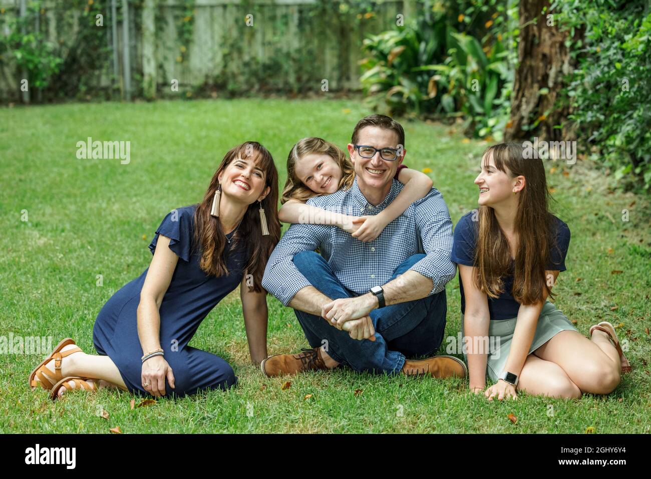 Eine Familie aus Mutter, Vater und zwei Töchtern, die draußen im Gras ihres Hinterhofs sitzen, umarmt die jüngste Tochter Vater von hinten Stockfoto