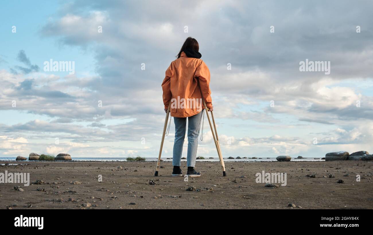 Eine Frau auf Krücken läuft am Ufer des Sees entlang. Sandstrand vor dem Hintergrund eines ruhigen Sees und Wolken am Horizont Stockfoto