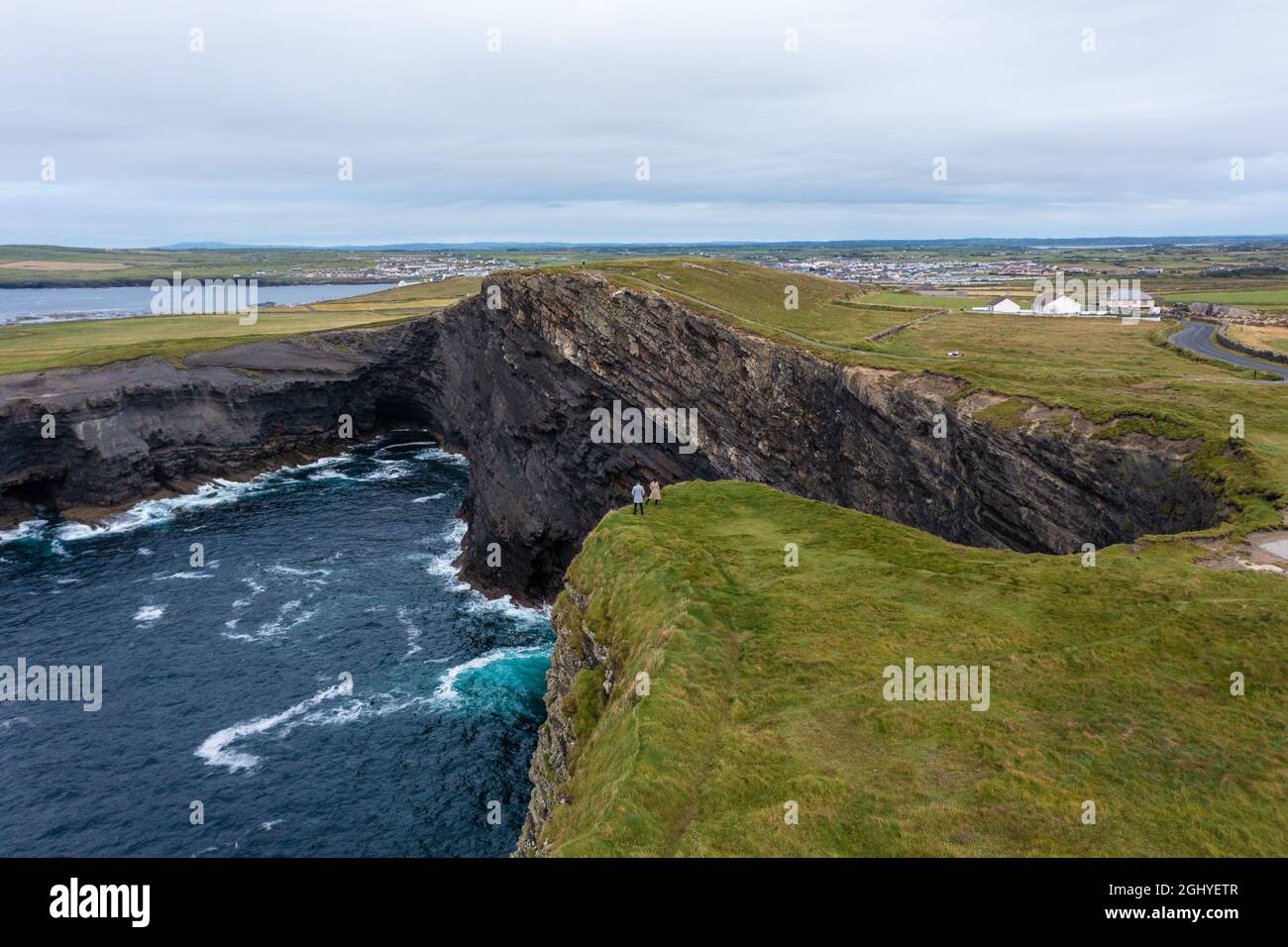 Luftaufnahme von nicht erkannten Menschen, die am Rand des Moher Cliff in Irland stehen, mit Meereswellen, die tagsüber unter bewölktem Himmel auf Felsen treffen Stockfoto