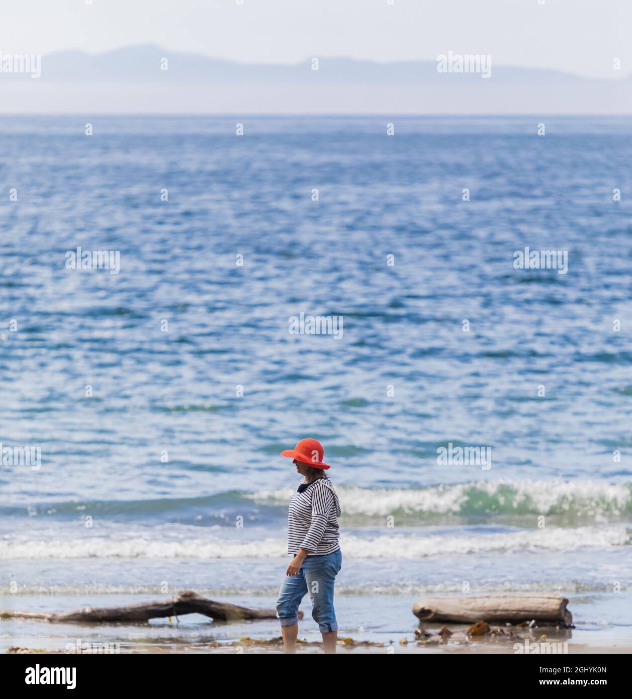 Eine Frau mit rotem Hut, die allein am leeren Strand am Ufer des Meeres läuft. Vancouver Island, BC, Kanada. August 9,2021. Straßenansicht, Reisefoto, Kopierbereich, sele Stockfoto