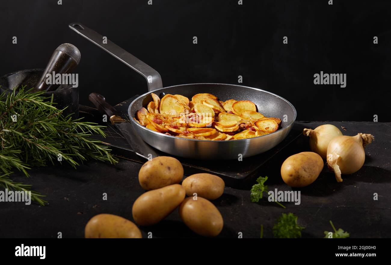 Pfanne mit appetitlich gerösteten Kartoffeln serviert auf schwarzem Tisch mit Zwiebeln und Rosmarin Zweige Stockfoto