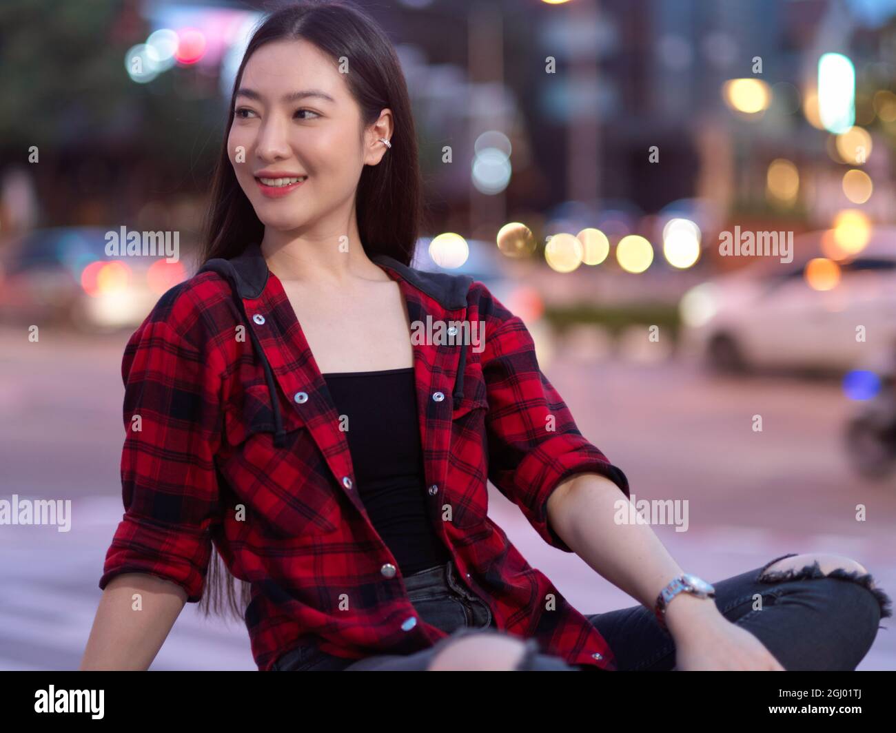 Attraktive, elegante junge Frau, die lächelt und von der Kamera wegschaut, sitzt auf der Stadtstraße, verwackelt im Straßennachtlicht im Hintergrund Stockfoto