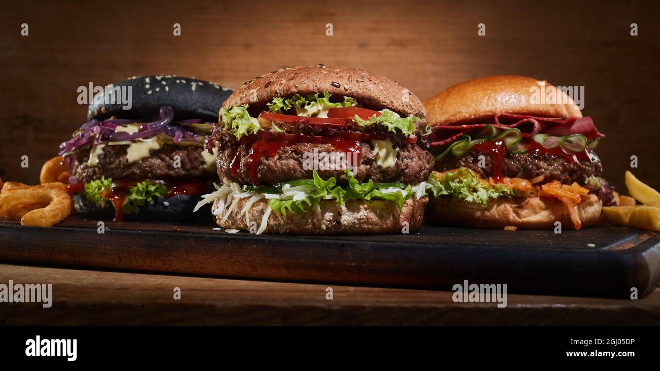 Appetitliche Hamburger mit Koteletts und verschiedenen Gemüsesorten, serviert auf Holzschneidebrett im Studio Stockfoto
