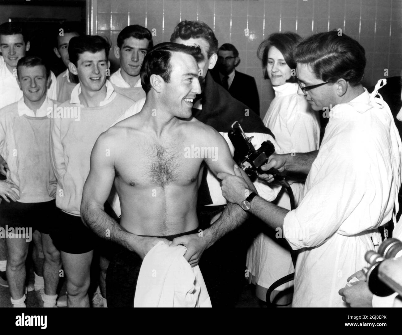 Der englische Stürmer Jimmy Greaves, der von seinen Teamkollegen aus Tottenham beobachtet wird, hat seine Anti-Influenza-Impfung. Oktober 1964. Stockfoto