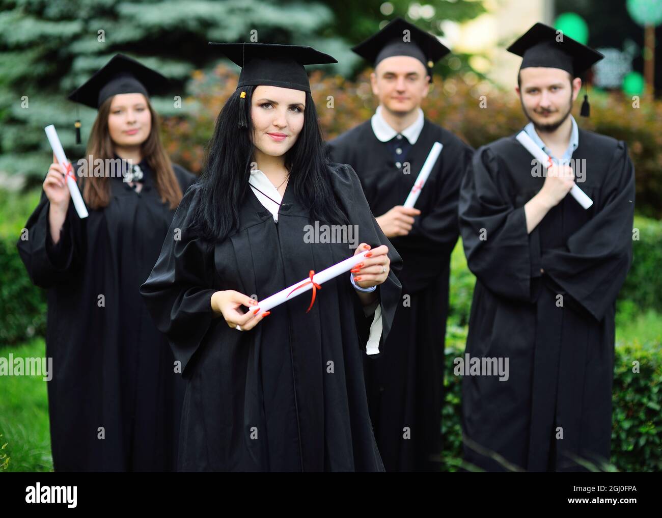Das junge Mädchen, eine Universitätsstudentin in einem schwarzen Gewand und einem quadratischen Hut mit einem Diplom in der Hand, lächelt vor dem Hintergrund der Studenten Stockfoto