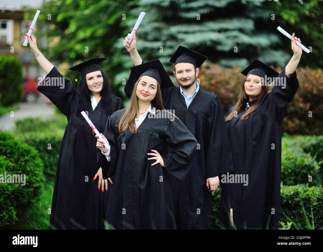 Das hübsche junge Mädchen ist eine Studentin in einem schwarzen Gewand und lächelt und hält vor dem Hintergrund einer Gruppe von Absolventen ein Diplom in den Händen. Stockfoto