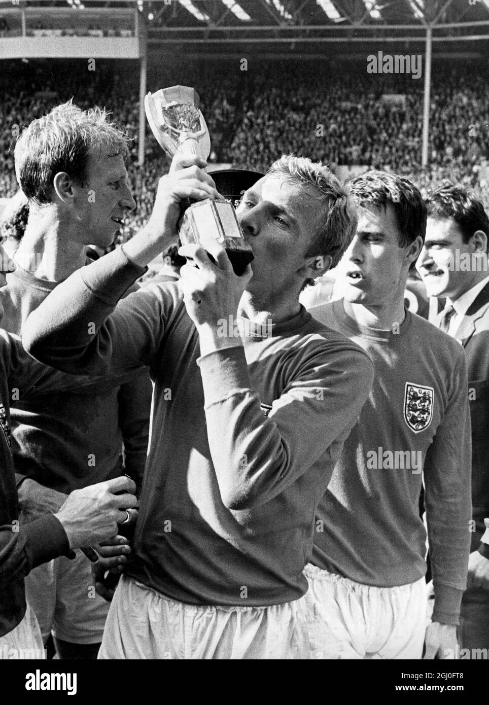 Der englische Kapitän Bobby Moore lässt sich begeistert von der Jules Rimet Trophy küssen, die ihm die Königin überreichte, nachdem er seine Männer im Weltcup-Finale im Wembley-Stadion (nach mehr Zeit) zum 4-2. Sieg über Westdeutschland geführt hatte. Auf der rechten Seite ist Geoff Hurst, der drei der England Tore erzielte und auf der linken Seite ist Jack Charlton. 30. Juli 1966 Stockfoto