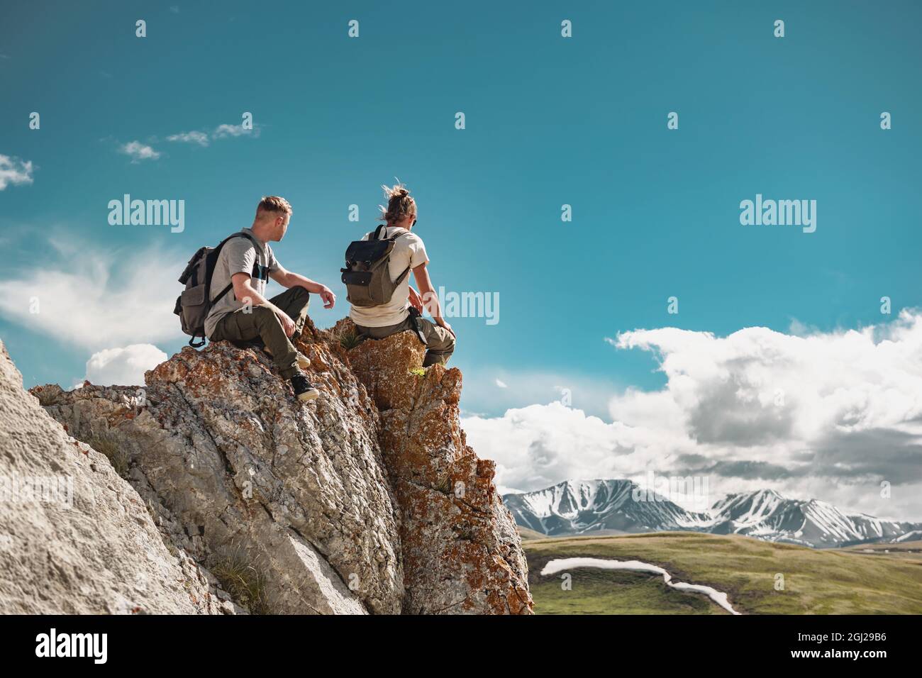 Zwei junge Wanderer mit kleinen Rucksäcken entspannen sich auf einem großen Felsen oder einer Klippe und genießen den Blick auf die Berge Stockfoto