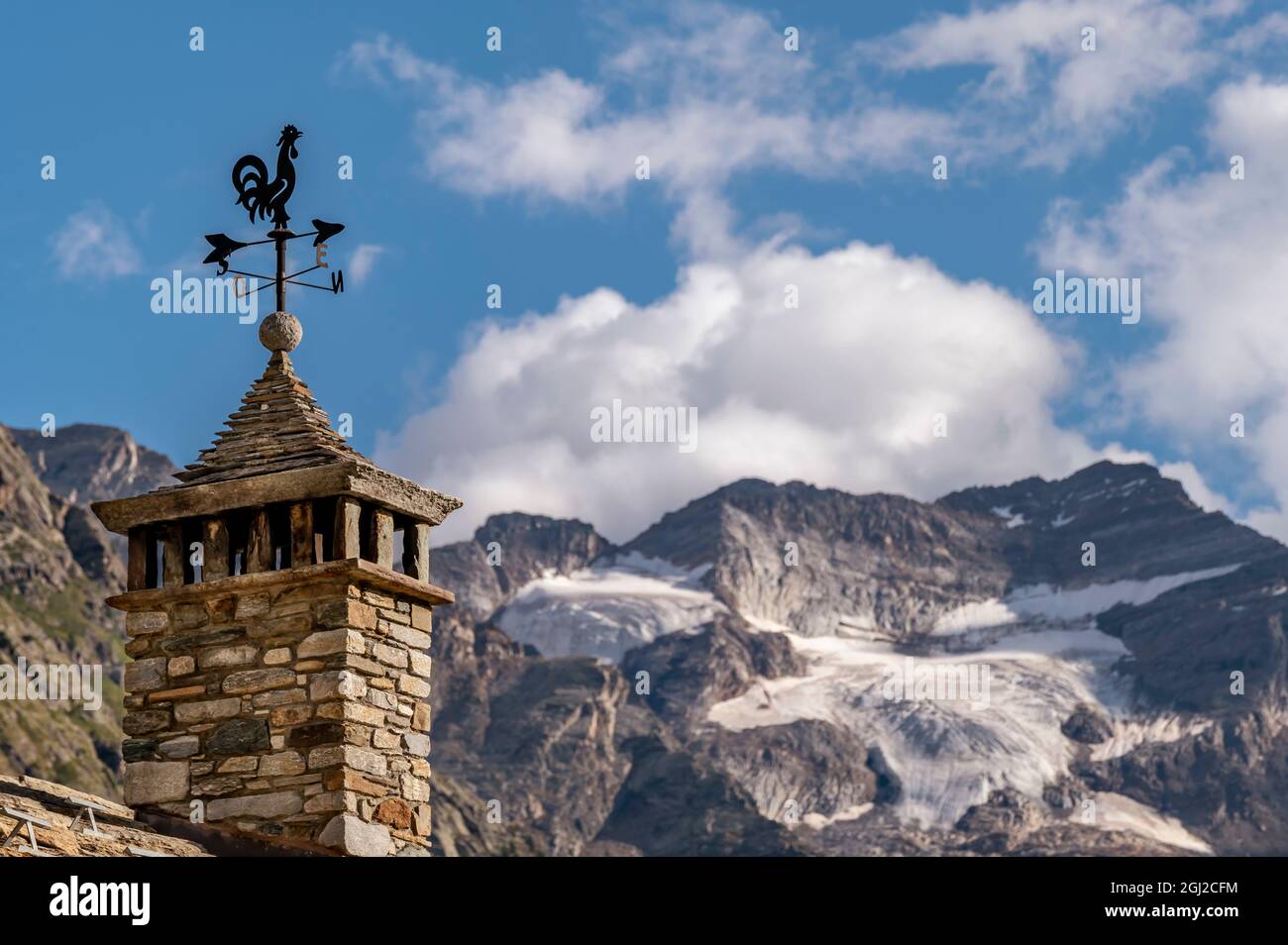 Eine Hahn-förmige Wetterfahne auf einem Schornstein in Lillaz, Aostatal, Italien, mit dem Gran Paradiso Gletscher im Hintergrund Stockfoto