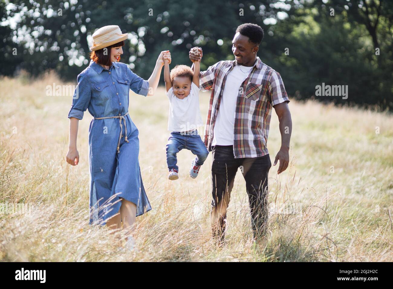 Fröhliche junge Eltern, die ihren Sohn für beide Hände aufziehen, während sie zusammen auf dem Sommerfeld spielen. Multirassische Familie in legerem Outfit, die jeden Moment zusammen verbringt. Stockfoto