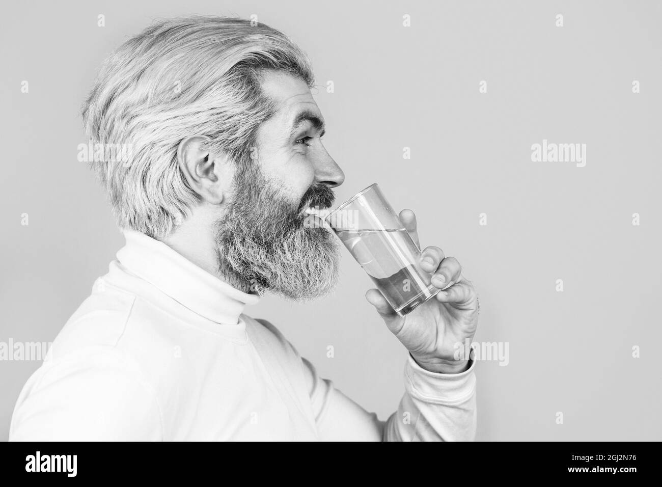 Lächelnder Mann mit transparentem Glas in der Hand. Bärtiger Mann, der Wasser trinkt. Mann trinkt aus einem Glas Wasser. Glücklicher Bartmann, der Wasser trinkt Stockfoto