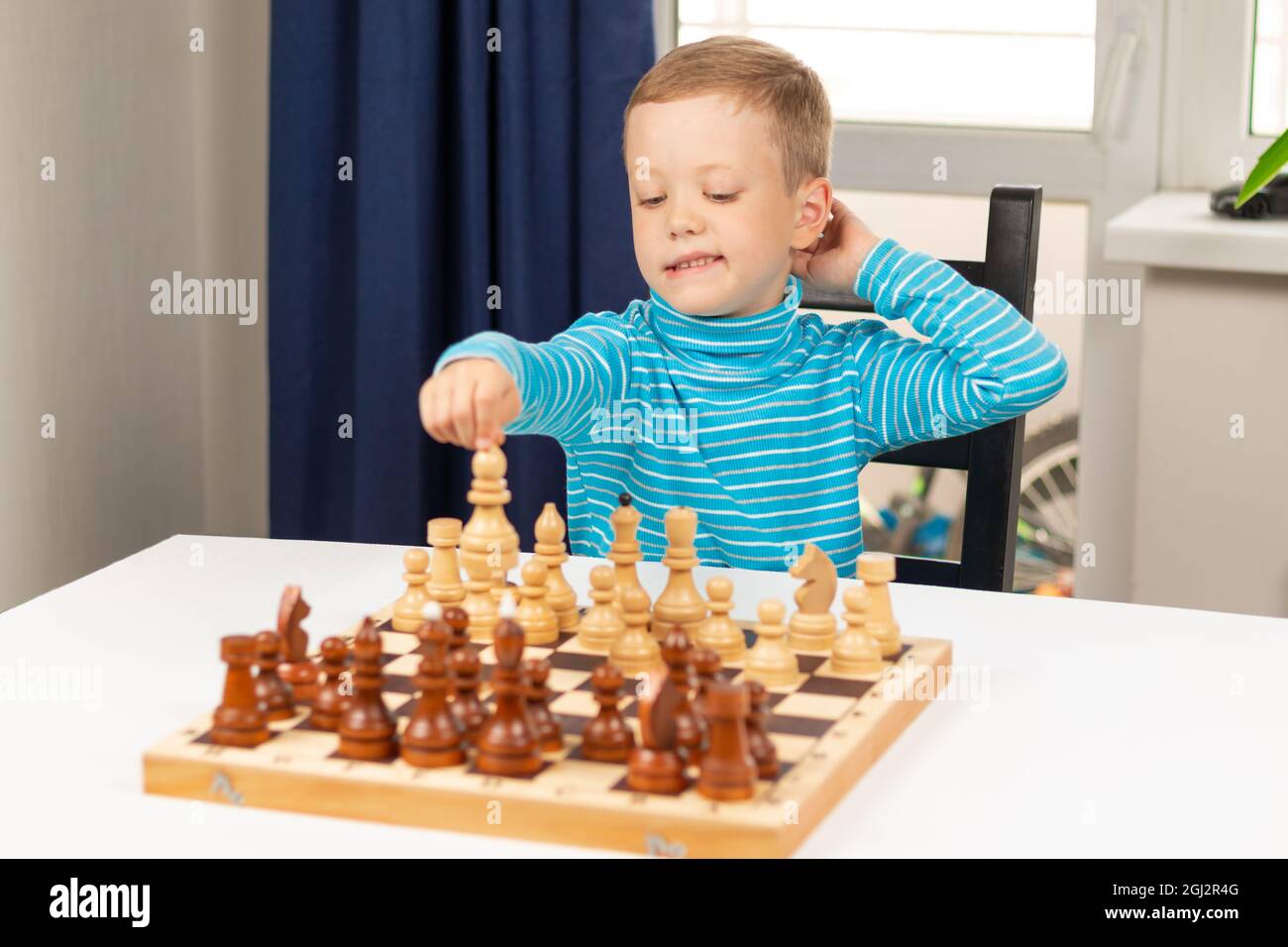 Netter siebenjähriger Junge, der zu Hause auf einem weißen Holztisch Schach spielt. Selektiver Fokus. Nahaufnahme. Hochformat Stockfoto