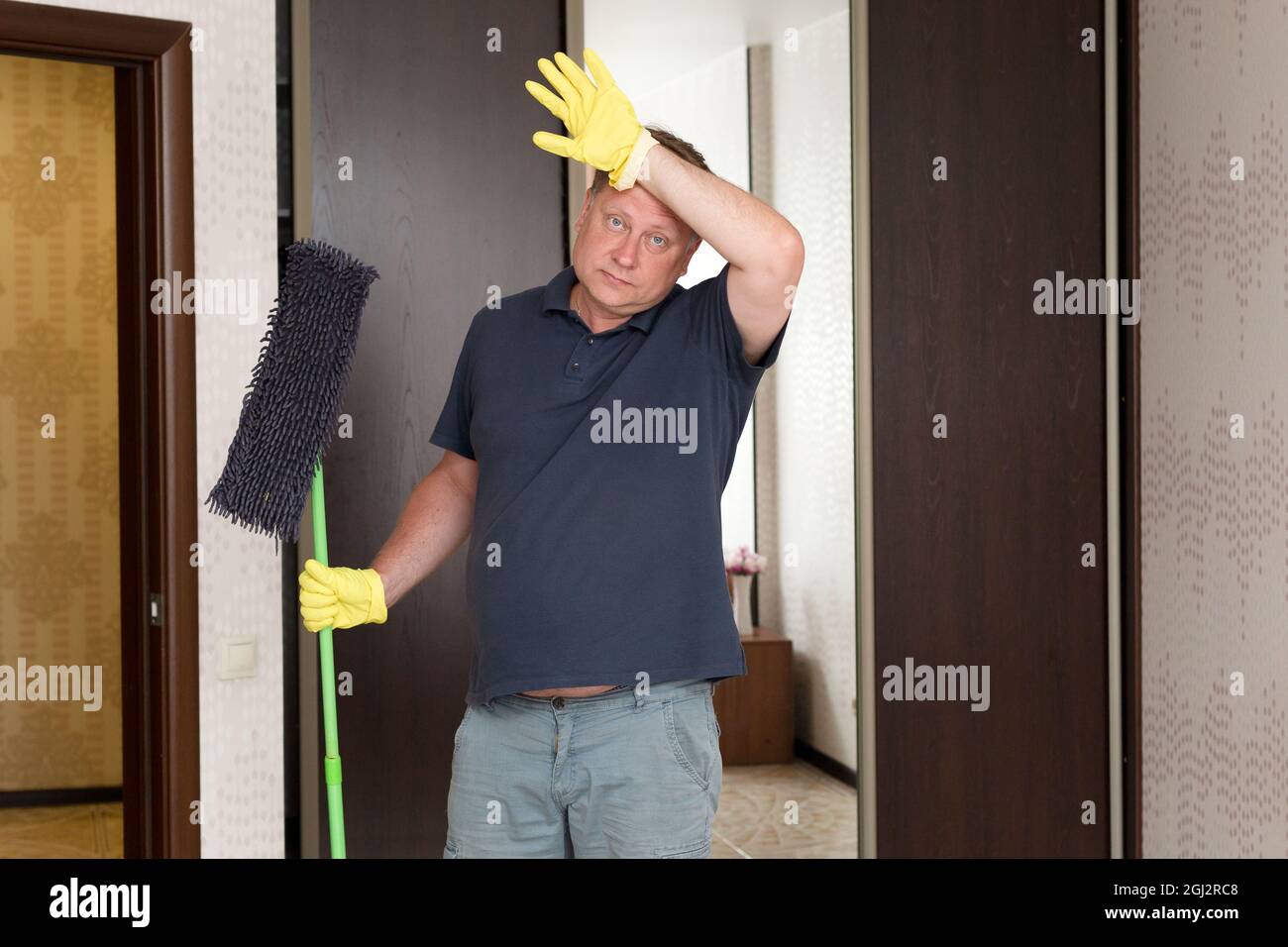 Ein Mann in gelben Gummihandschuhen mit einem Wischmopp in den Händen reibt sich nach der Reinigung der Wohnung vor Müdigkeit die Stirn. Stockfoto