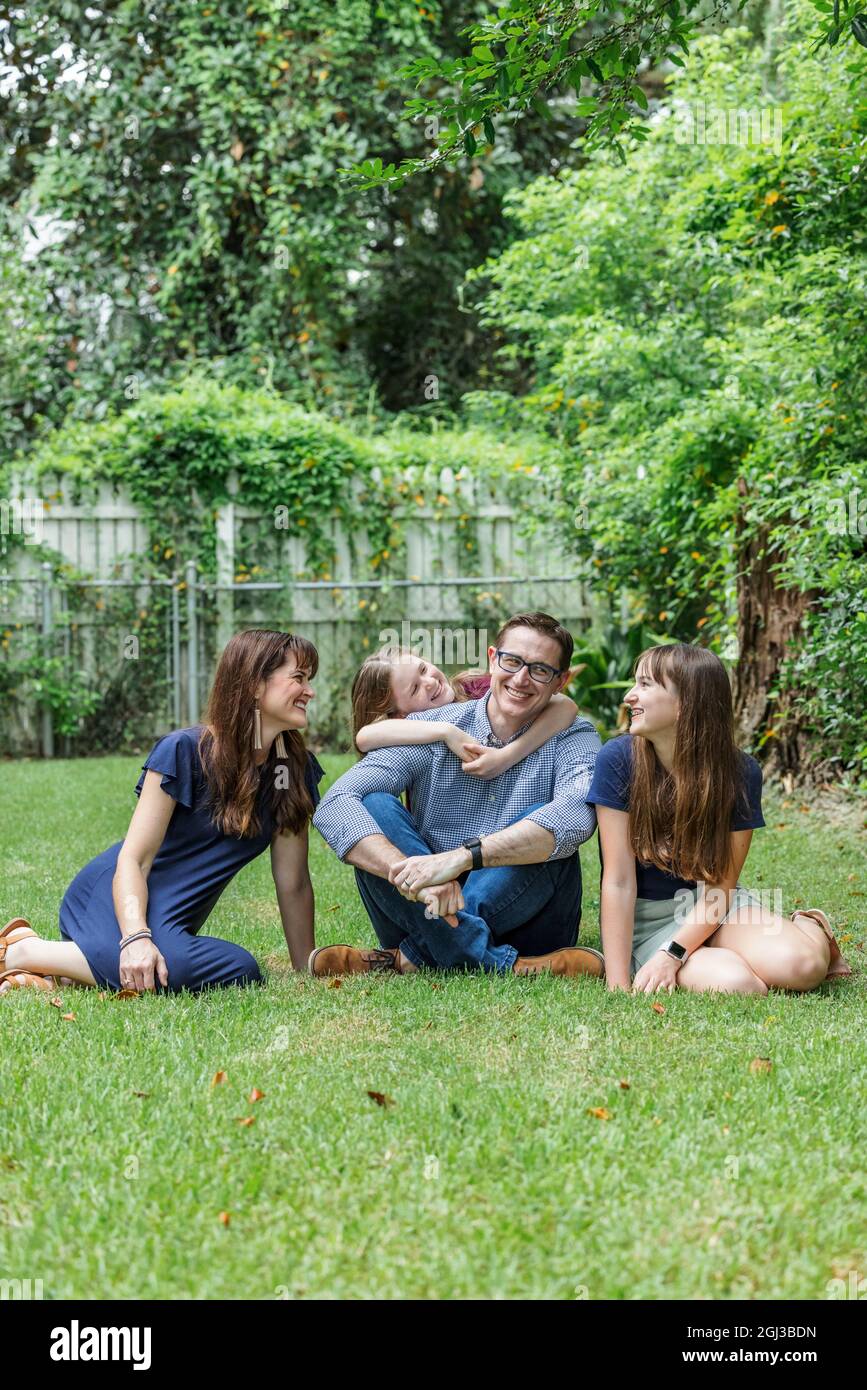 Eine Familie aus Mutter, Vater und zwei Töchtern, die draußen im Gras ihres Hinterhofs sitzen, umarmt die jüngste Tochter Vater von hinten Stockfoto