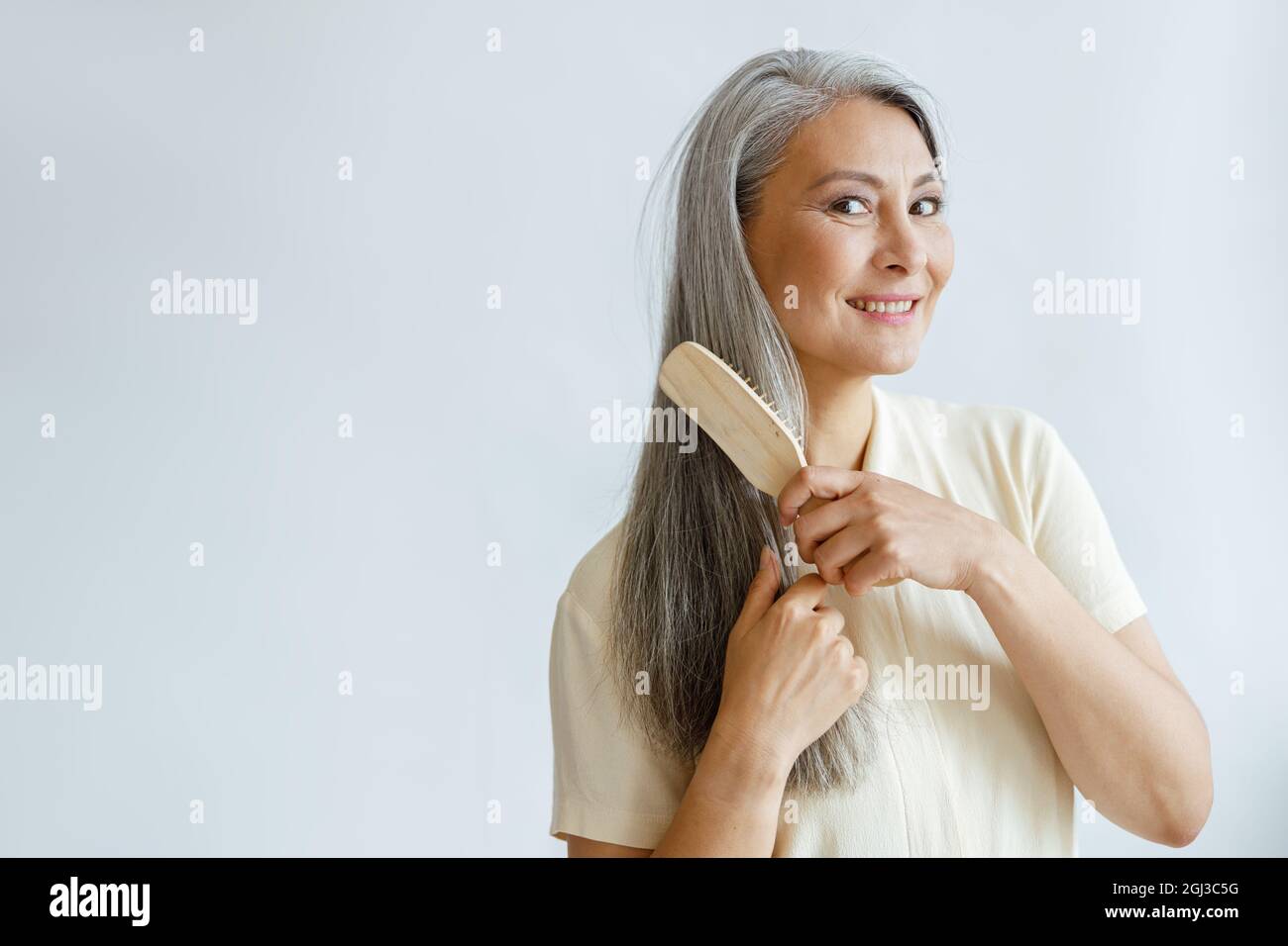 Nachdenkliche asiatische Frau mittleren Alters bürstet graue Haare posiert auf hellem Hintergrund Stockfoto