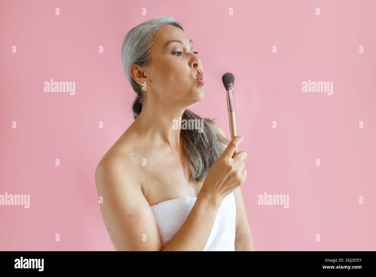 Lustige asiatische Dame mittleren Alters mit weißem Handtuch umwickelt bläst kosmetischen Pinsel auf rosa Hintergrund Stockfoto