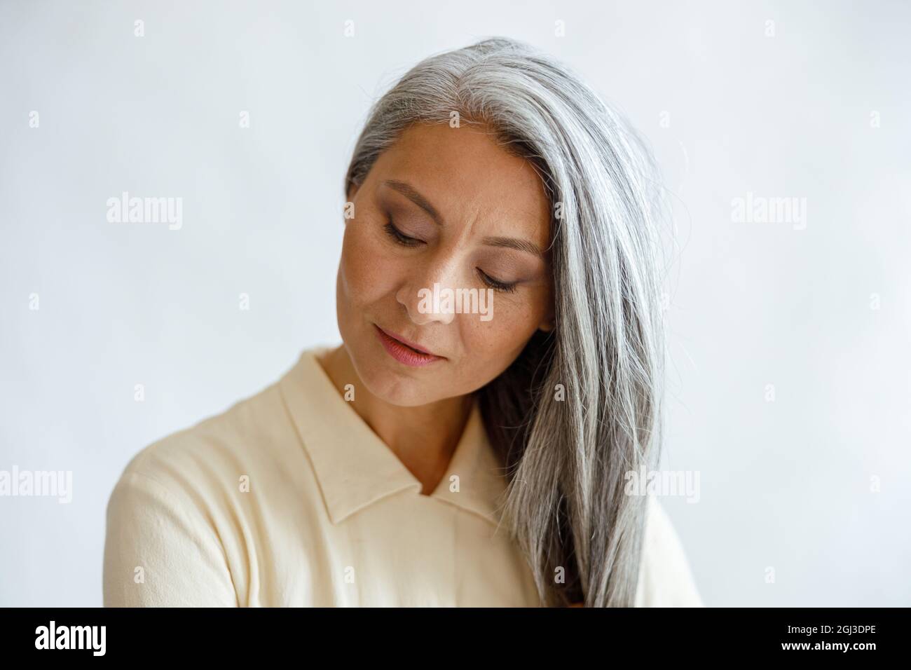 Ruhige asiatische Frau mittleren Alters mit lockeren silbernen Haaren, die auf hellem Hintergrund posiert Stockfoto