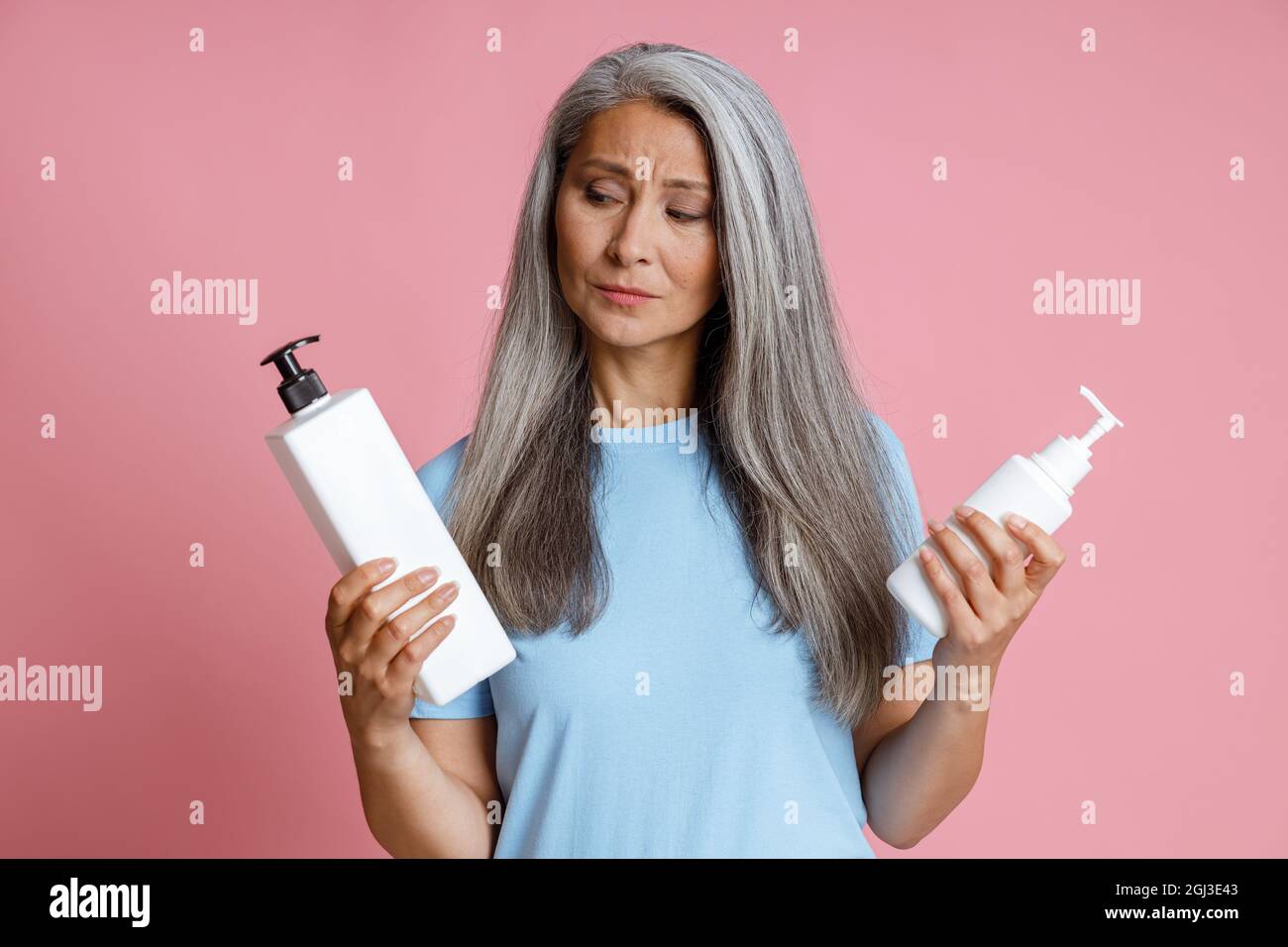 Nachdenkliche asiatische Dame mittleren Alters wählt kosmetisches Produkt auf rosa Hintergrund Stockfoto