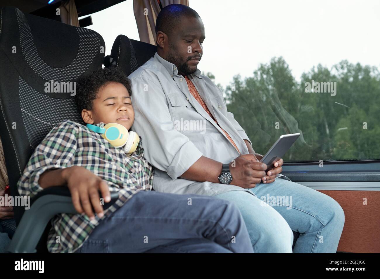 Afroamerikanischer Junge mit Kopfhörern um den Hals, der im Bus auf der Schulter des Vaters schläft, während er ein Tablet benutzt Stockfoto