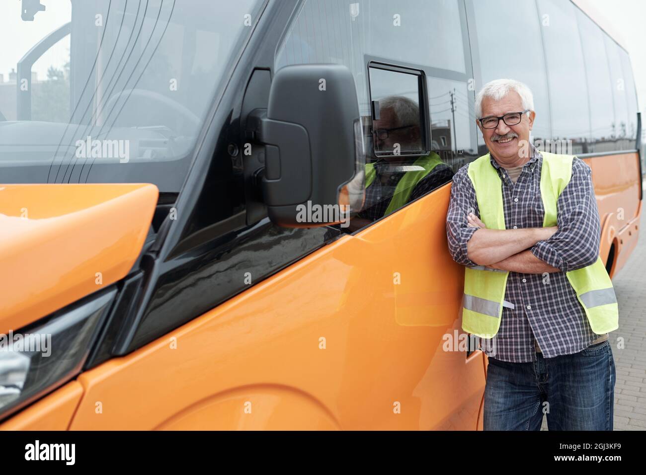 Porträt eines lächelnden weißhaarigen älteren Busfahrers in einer Brille, der mit gekreuzten Armen steht und sich auf einen orangefarbenen Bus stützt Stockfoto