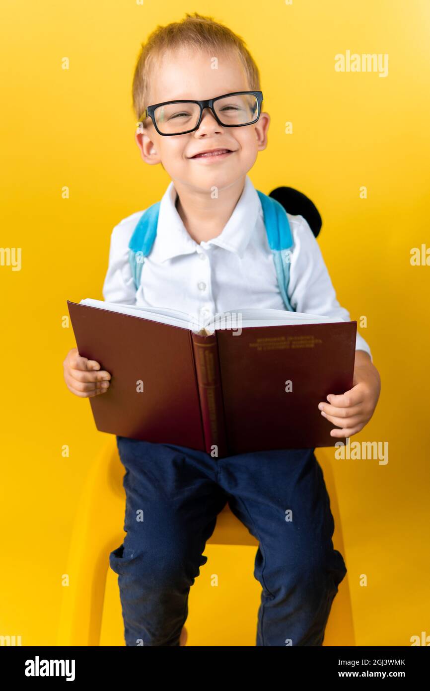 Porträt von Lustige Vorschule Kind Junge in großen Gläsern sitzt auf Stuhl liest Buch auf gelbem Hintergrund. Happy Smiling Kid Gehen Sie zurück zur Schule, Kindergarten Stockfoto