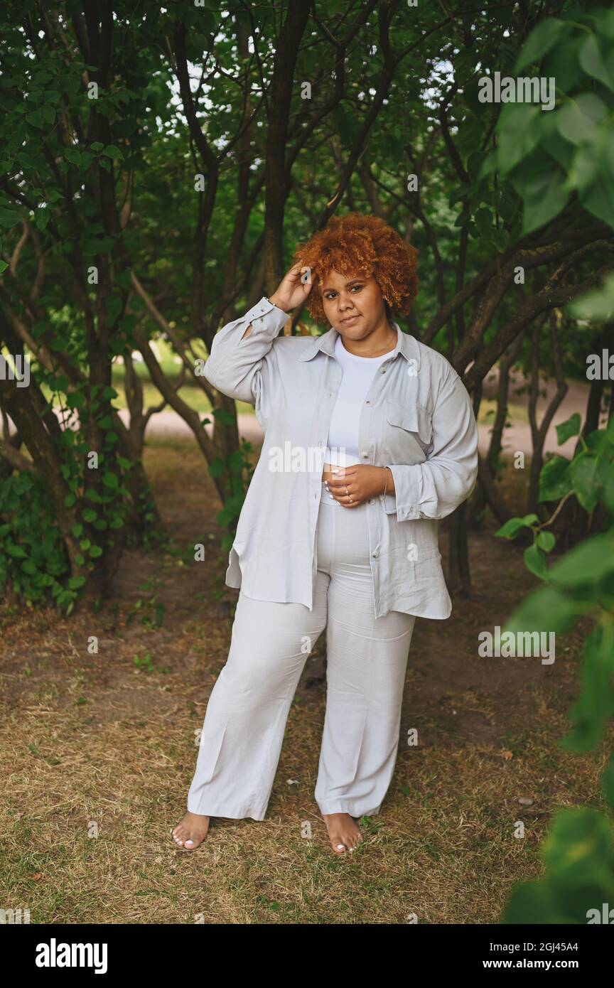 Fashion Portrait von jungen natürlich schönen plus size afroamerikanische Frau in hellen Hosenanzug mit afroroten Haaren posiert in Natur Parkland in grünem Laub. Vielfalt und körperpositives Konzept Stockfoto
