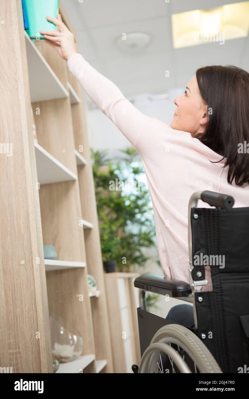 Frau im Rollstuhl, die aus dem hohen Regal nach Büchern greift Stockfoto