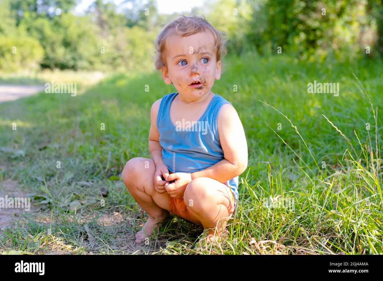 Überrascht hockt der kleine Junge auf dem Gras. Das Gesicht des Jungen ist mit Schmutz verschmiert Stockfoto