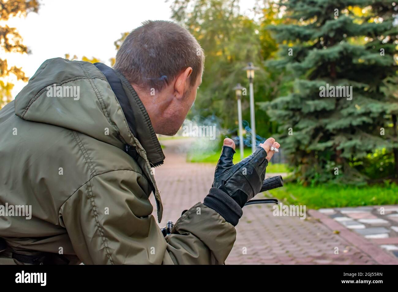 Die Hand eines nachdenklichen Mannes hält eine rauchende Zigarette, die auf der Straße der Stadtallee des Parks steht, ein Blick von hinten, hinten Stockfoto