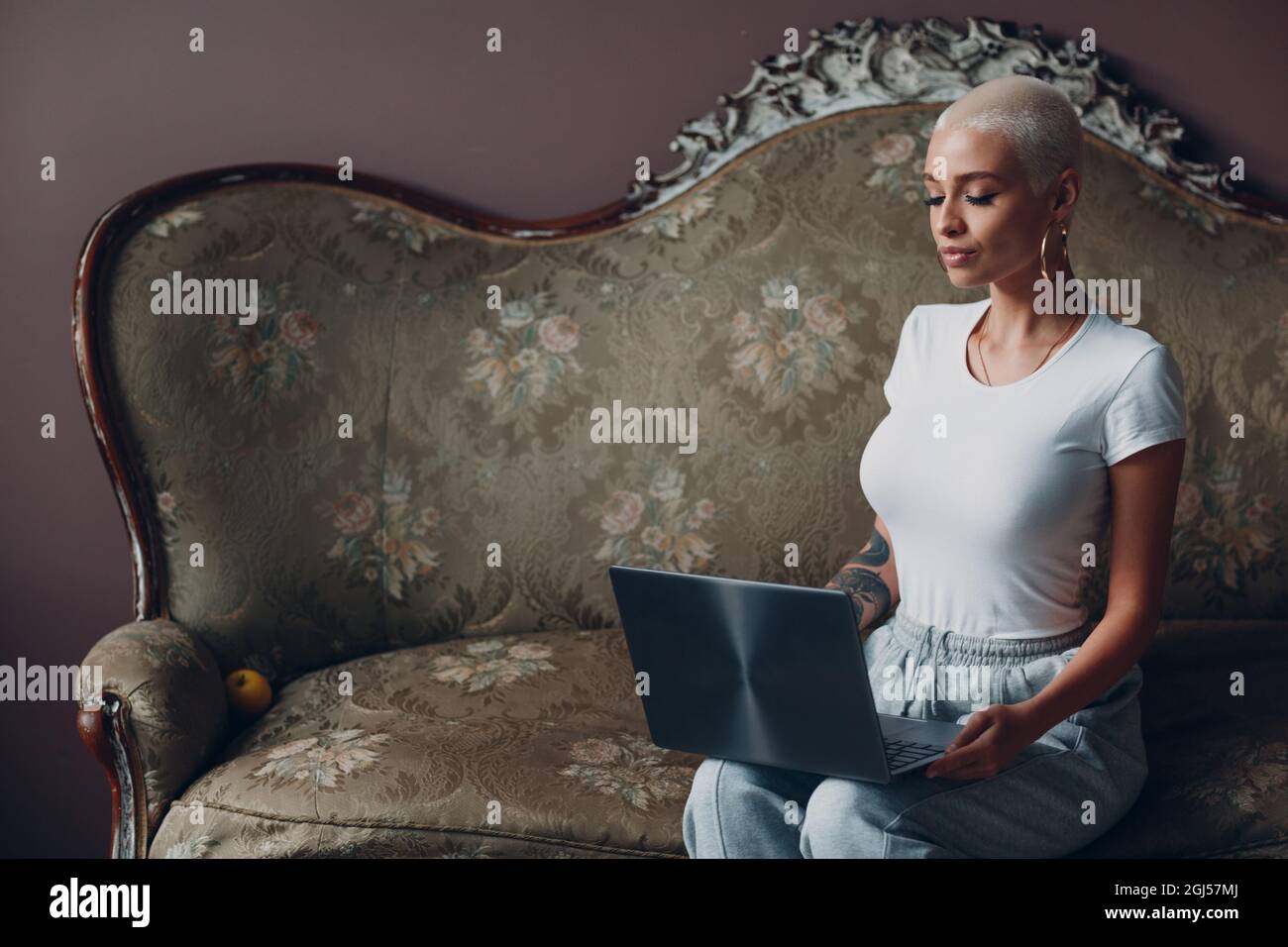 Eine junge Frau mit kurzen blonden Haaren, die mit einem Laptop auf einem Vintage-Sofa im Home Office sitzt Stockfoto