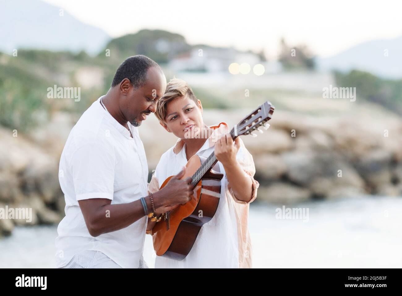Fröhliches gemischtes Rennen, Paar mittleren Alters, das sich umarmt, während man am Strand mit einer Gitarre läuft. Mann mit Gitarre und Frau. Stockfoto