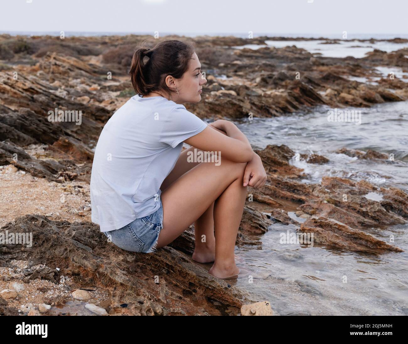 Nachdenkliches Teenager-Mädchen, das bei Sonnenuntergang auf einer Klippe am Meer sitzt, die Beine im Wasser hat und gerade aussieht, blaues T-Shirt und Jeans-Shorts trägt und Side Vie trägt Stockfoto