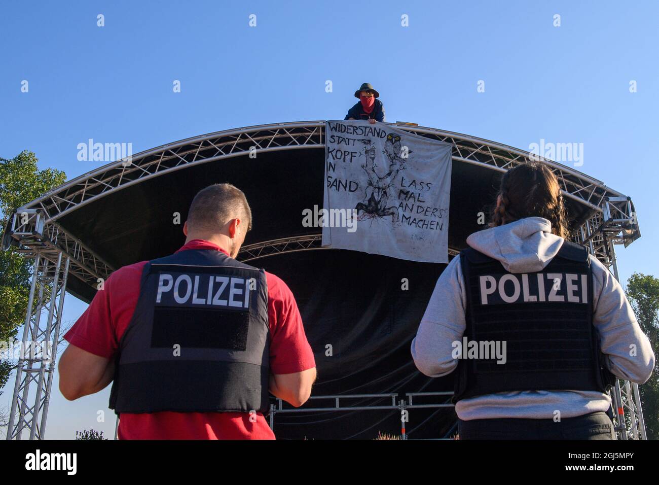 09. September 2021, Sachsen-Anhalt, Rochau: Polizisten stehen vor einem Autobahngegner, der auf dem Dach einer Bühne sitzt, die für den feierlichen Baubeginn der Autobahnumgehung A14 in Stendal eingerichtet wurde. Sein Schal lautet: „besser widerstehen, als den Kopf in Sand zu stecken“ und „Let's do things differently“. Bei der Veranstaltung war mit dem Bundesverkehrsminister und dem Verkehrsminister des Landes Sachsen-Anhalt ein Baum gepflanzt worden. Neben den Autobahngegnern waren auch Mitglieder von Initiativen, die den Autobahnbau begrüßten, am Standort. Foto: Klaus-Dietma Stockfoto