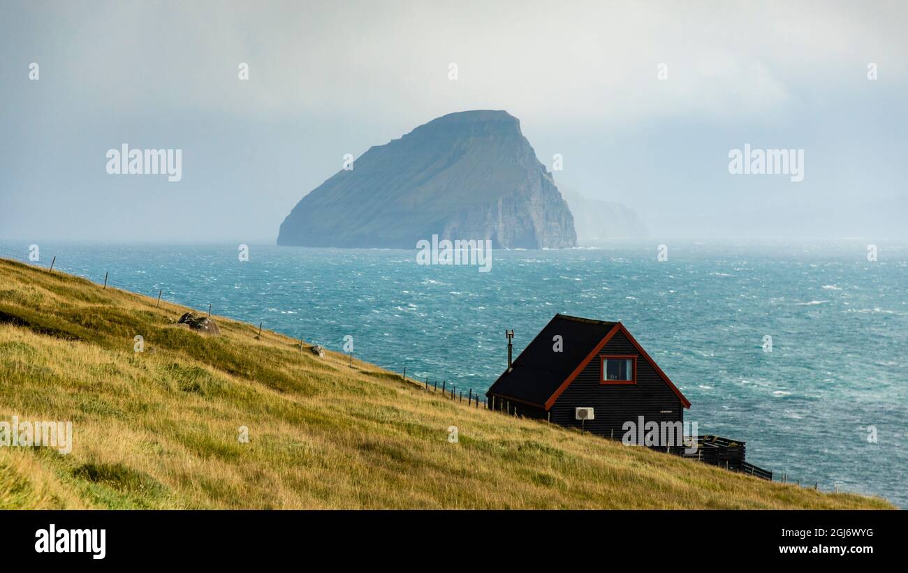 Europa, Färöer. Blick auf das traditionelle Haus auf einer Klippe am Meer auf der Insel Streymoy, mit der Insel Kuttur im Hintergrund. Stockfoto