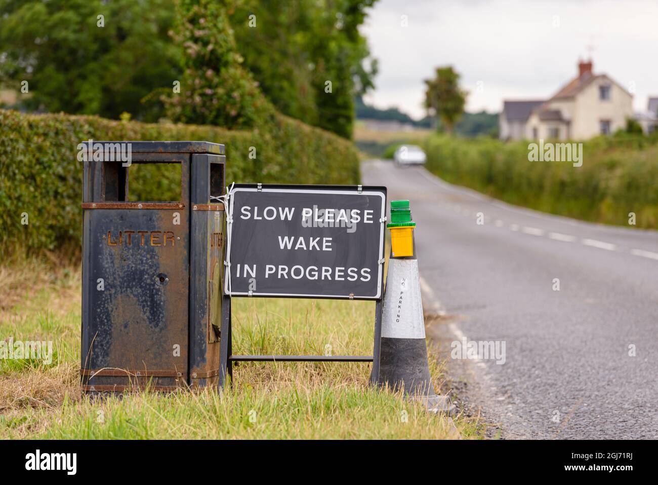 Zeichen mit einem schwarzen Hintergrund und den Worten "langsam bitte. Wake in Progress“ mit einem schwarz-weißen Verkehrskegel und einem Warnlicht an der Seite eines Roa Stockfoto