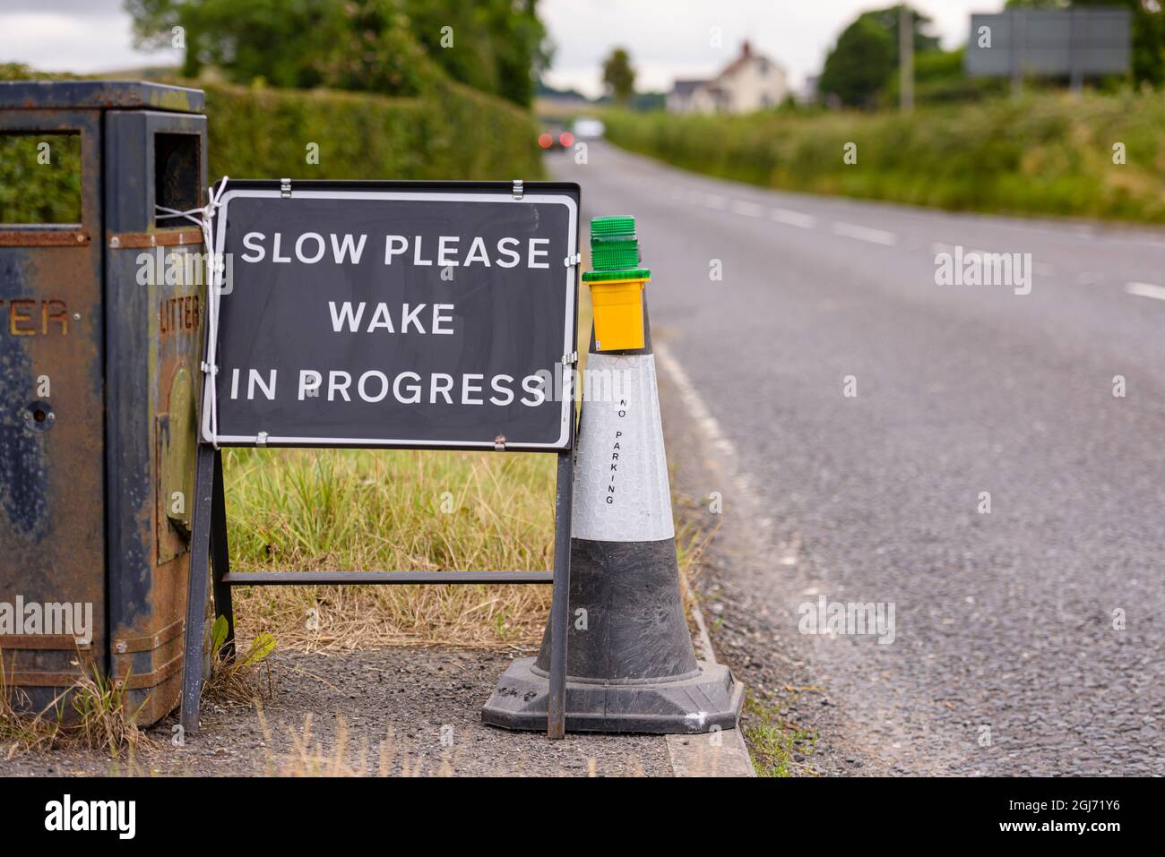 Zeichen mit einem schwarzen Hintergrund und den Worten "langsam bitte. Wake in Progress“ mit einem schwarz-weißen Verkehrskegel und einem Warnlicht an der Seite eines Roa Stockfoto
