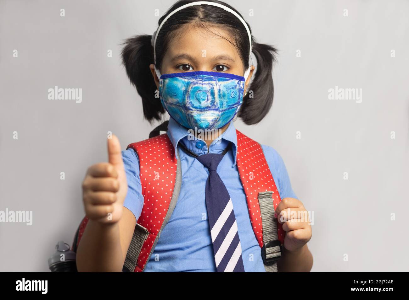 Ein fröhliches indisches Mädchen in blauer Schuluniform mit roter Tasche und Nasenmaskenschutz zeigt Daumen nach oben auf grauem Hintergrund Stockfoto