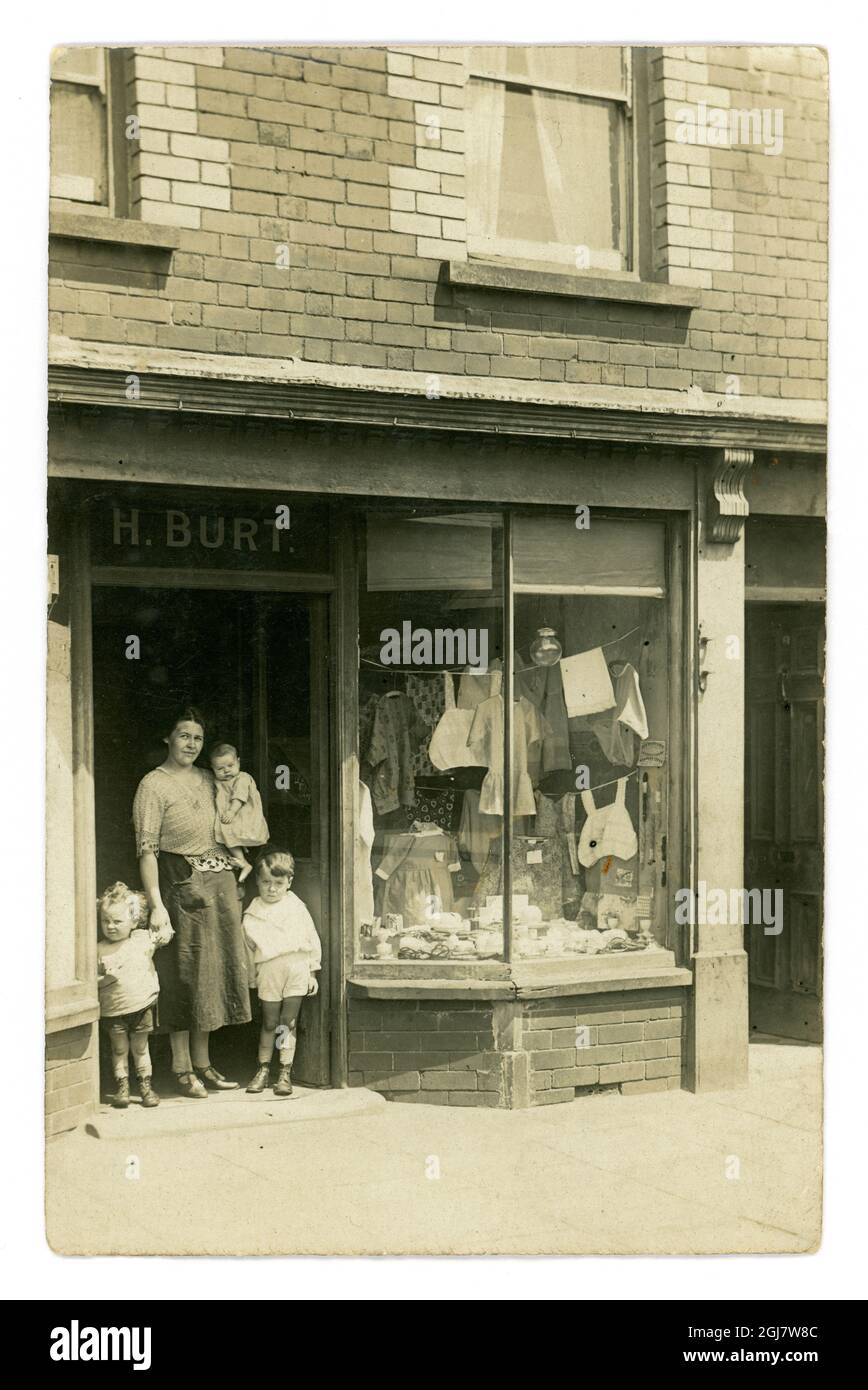 Original echte Fotopostkarte einer Arbeiterin mit ihren Kindern, die draußen im Ladeneingang eines Kurzwarenladens namens H. Burt stehen, der Wolle, Schürzen, Kamisolen verkauft, Wolle und Fäden. Die Frau sieht umwurmt aus und die Ladenfront ist schäbig, wahrscheinlich in einer armen Nachbarschaft, etwa 1921, Großbritannien Stockfoto