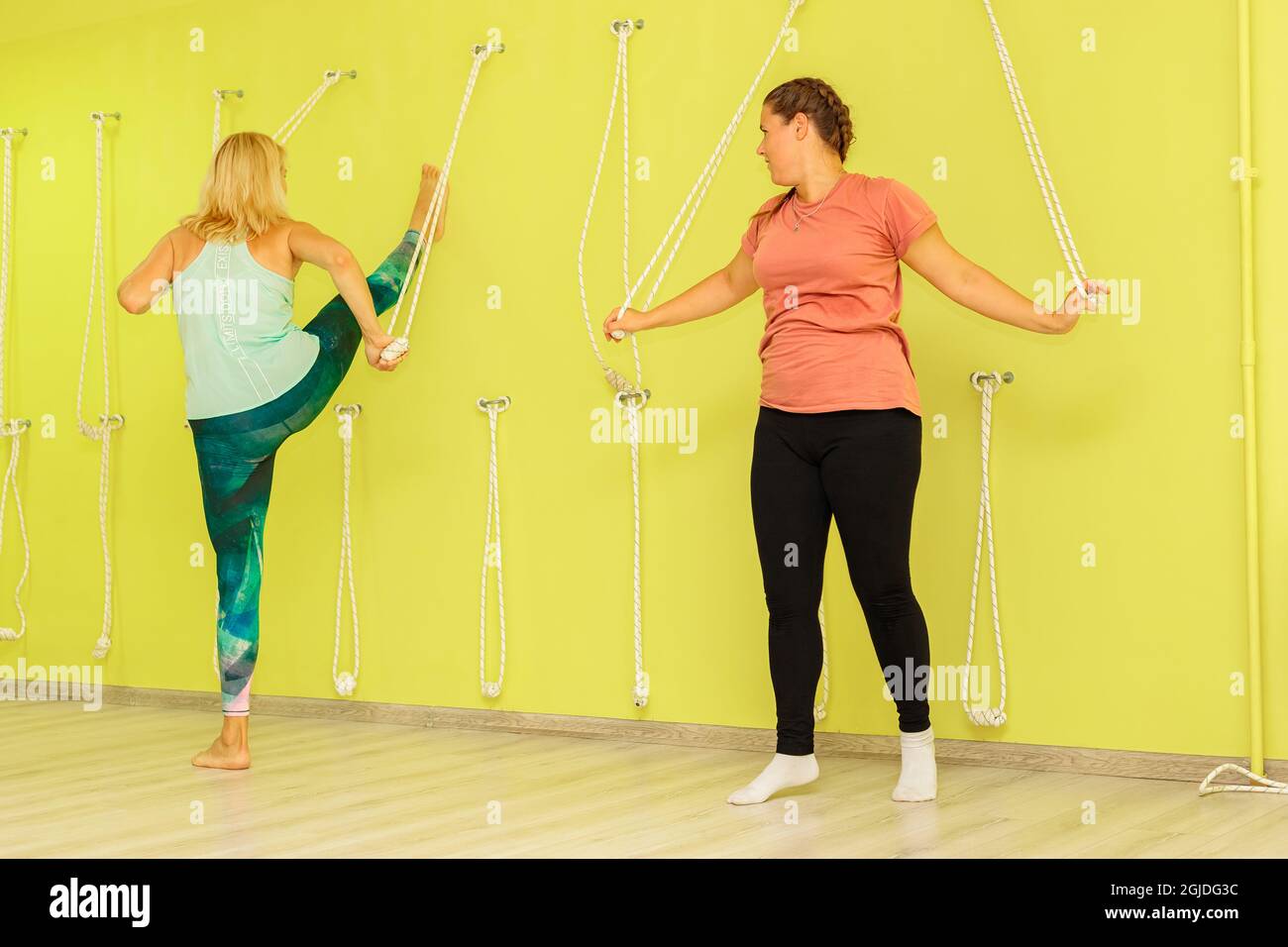 Ein Mädchen mit einem Ausbilder führt eine Übung in der Nähe einer gelben Wand mit Seilen im Fitnessstudio durch. Die Mädchen machen Yoga. Stockfoto