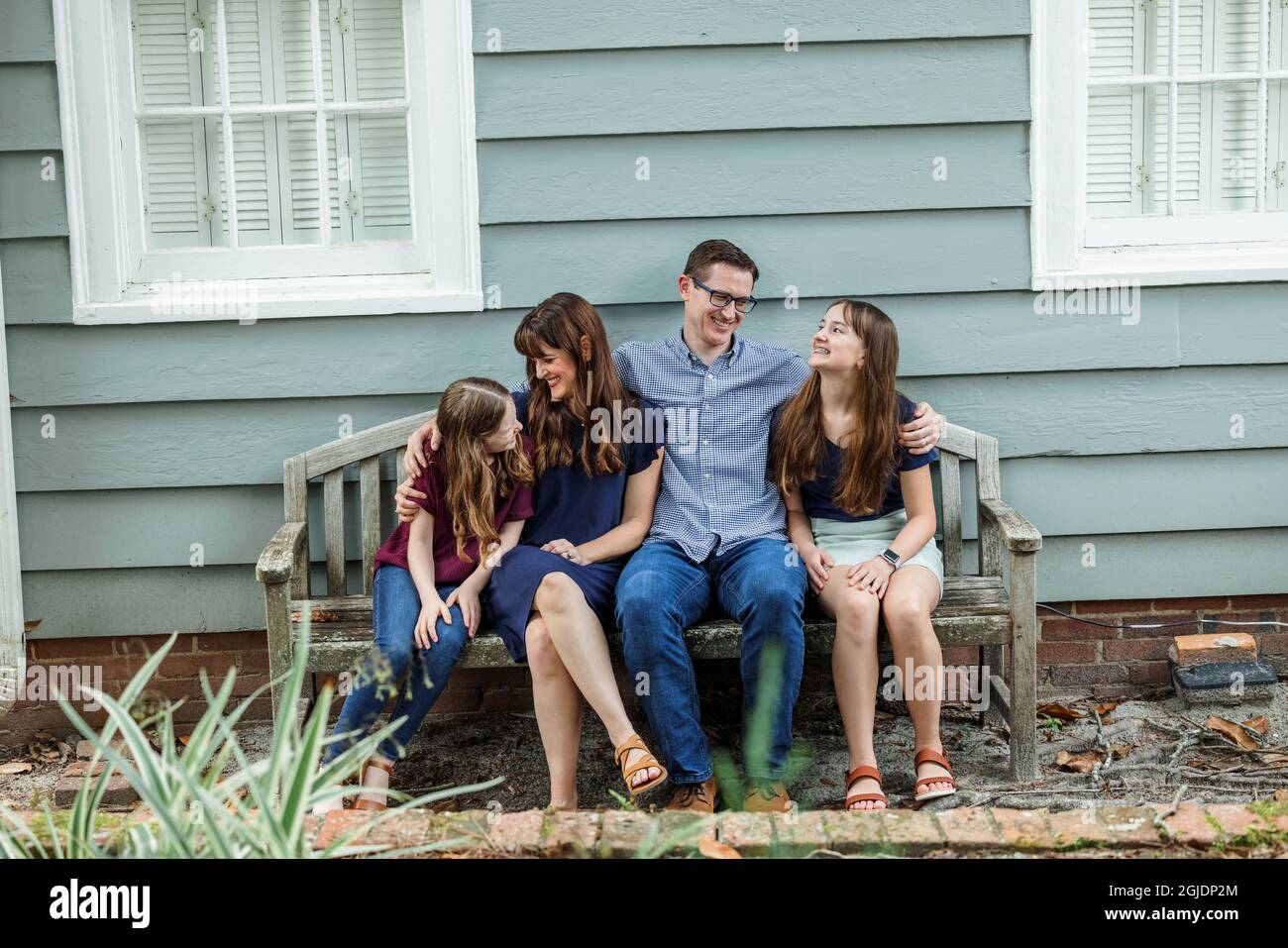 Eine vierköpfige Familie mit Mutter, Vater und zwei Töchtern, die auf einer Bank vor einem blauen Häuschen sitzen Stockfoto