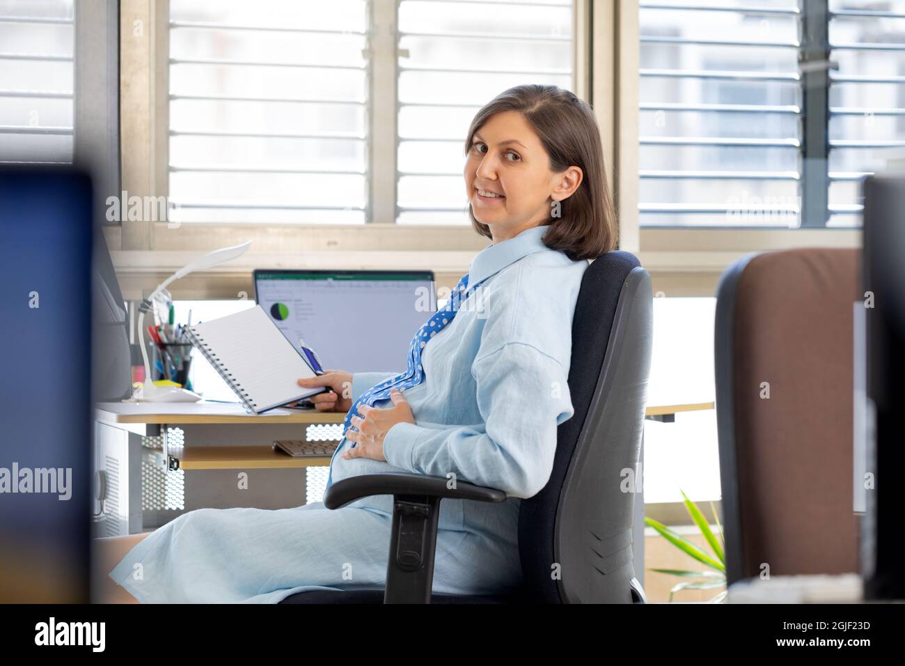 Eine Schwangerin arbeitet im Büro am Computer. Der Mitarbeiter sitzt auf einem Stuhl gegenüber dem Fenster und dreht sich um. Stockfoto