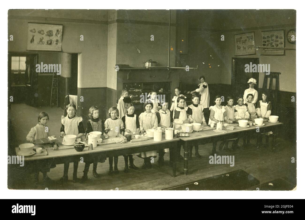 Original Postkarte aus dem frühen 20. Jahrhundert, Mädchen im Kochkurs, die an langen Tischen arbeiteten, Gemüse-, Fleisch- und Viehkarten an den Wänden, erstaunliche Backöfen, Brot und Scones backen - London, England, Großbritannien um 1903 Stockfoto