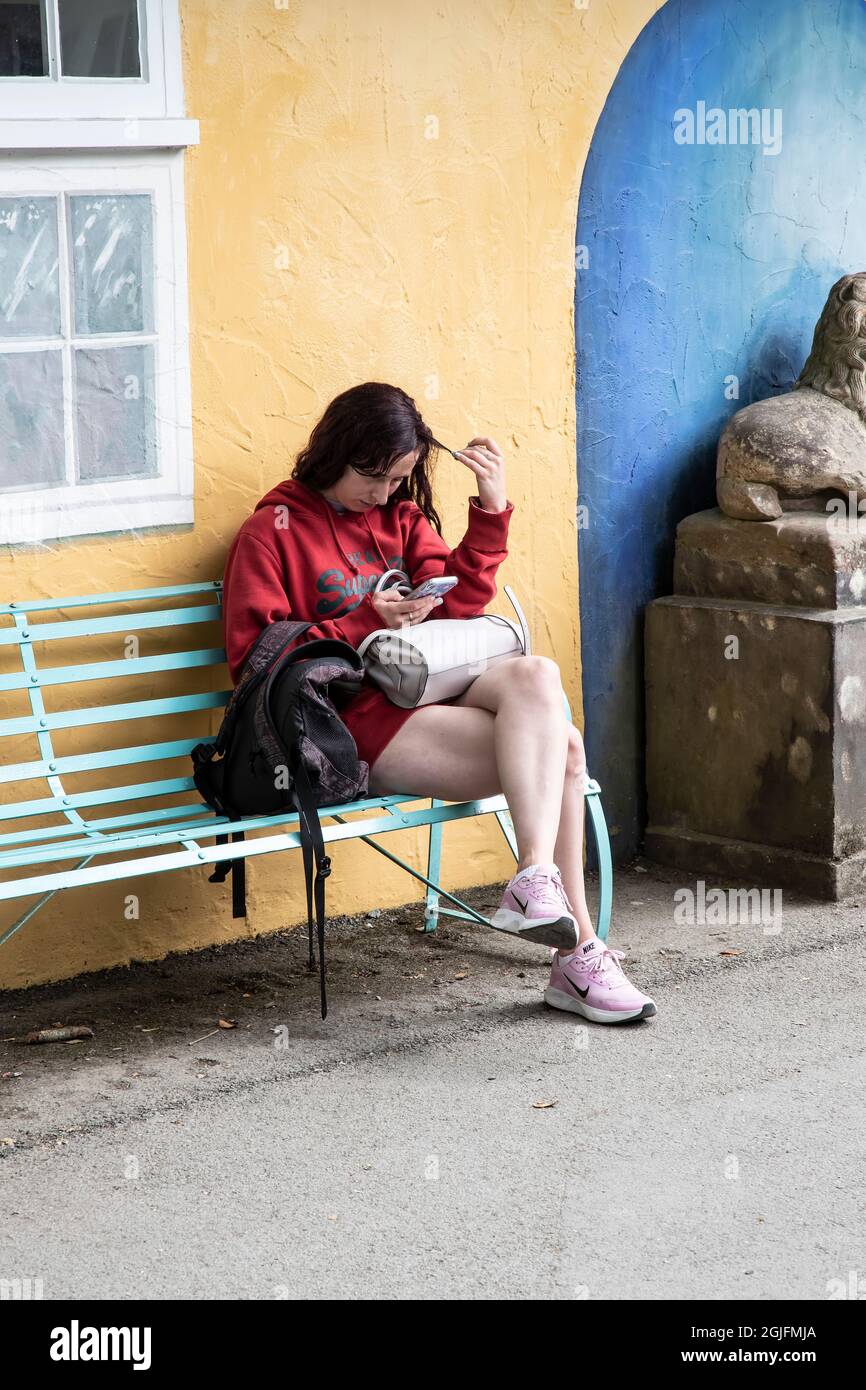 Das junge Mädchen mit Handtasche und Rucksack sitzt auf einer Bank, die mit ihren Haaren twiddling und beschäftigt ist, ein Mobiltelefon in Portmerion, Nordwales, zu lesen Stockfoto