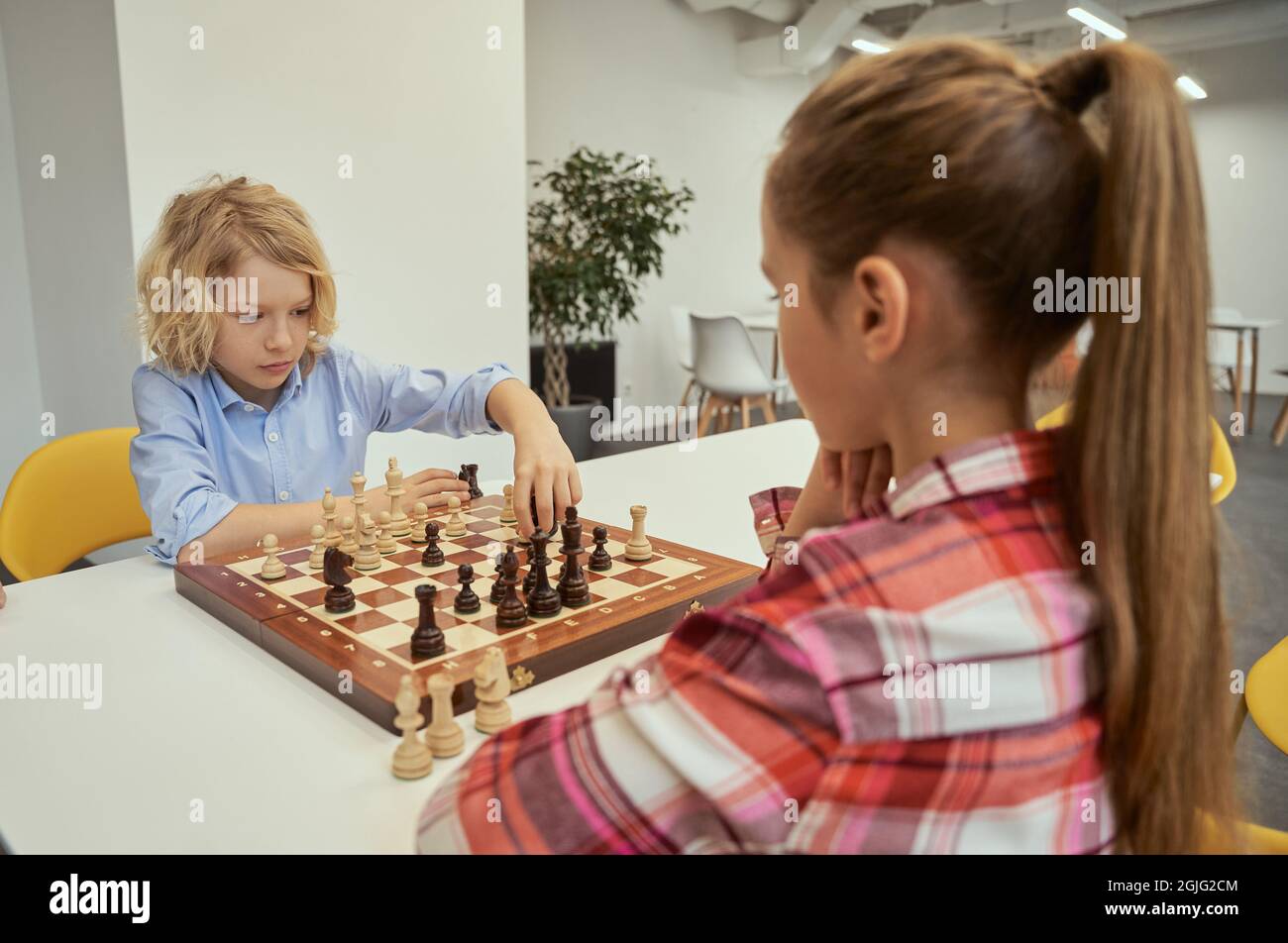 Ernsthafter kleiner kaukasischer Junge, der seinen Zug macht, während er mit einem Freund Schach spielt und zusammen an einem Tisch in der Schule sitzt Stockfoto