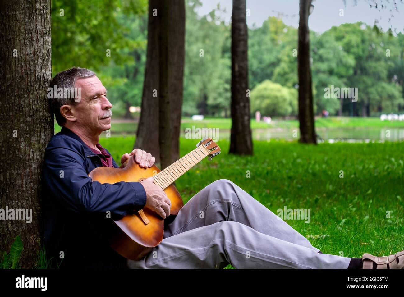 Am Sommerabend sitzt ein erwachsener Romantiker im schönen Park auf Gras, lehnt sich an einen Baum und spielt eine klassische 6-saitige Akustikgitarre. Selektiv Stockfoto