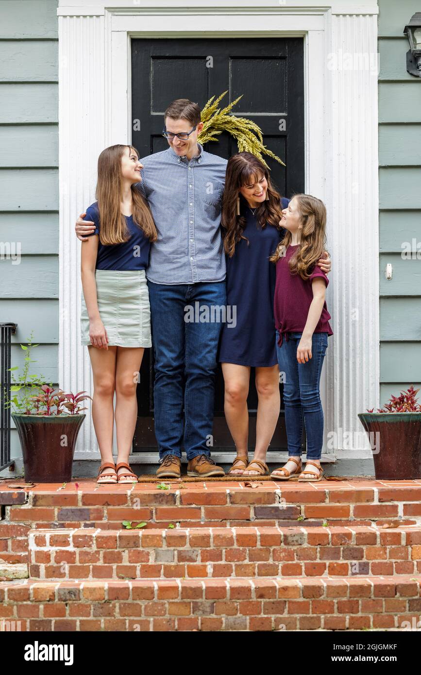 Eine Familie mit einer Mutter, einem Vater und zwei Töchtern, die draußen auf der Backsteinvertei eines kleinen blauen Häuschens stehen und sich gegenseitig betrachten Stockfoto