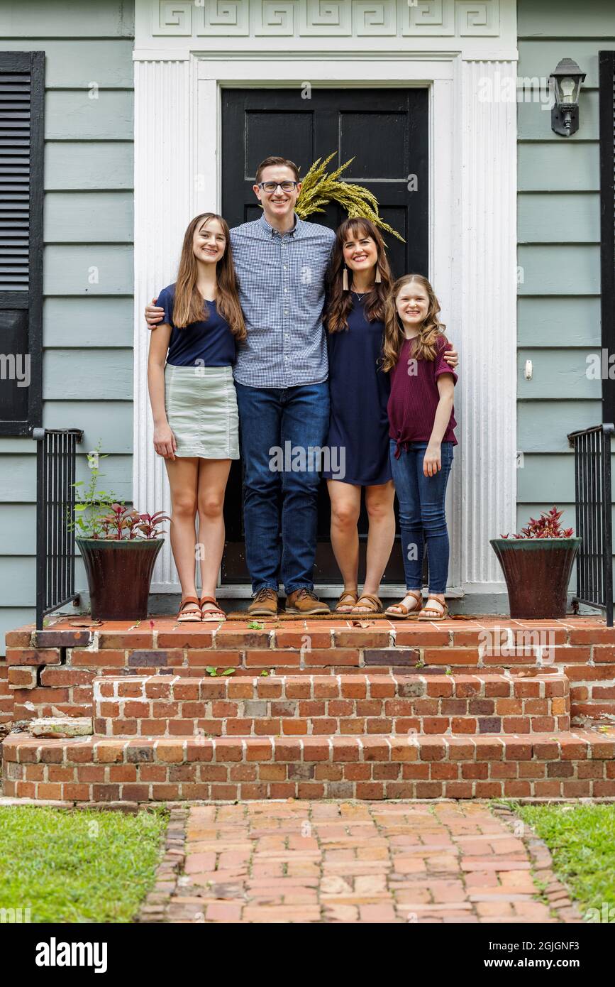 Eine Familie mit einer Mutter, einem Vater und zwei Töchtern, die draußen auf der Backsteinvertei eines kleinen blauen Häuschens stehen Stockfoto